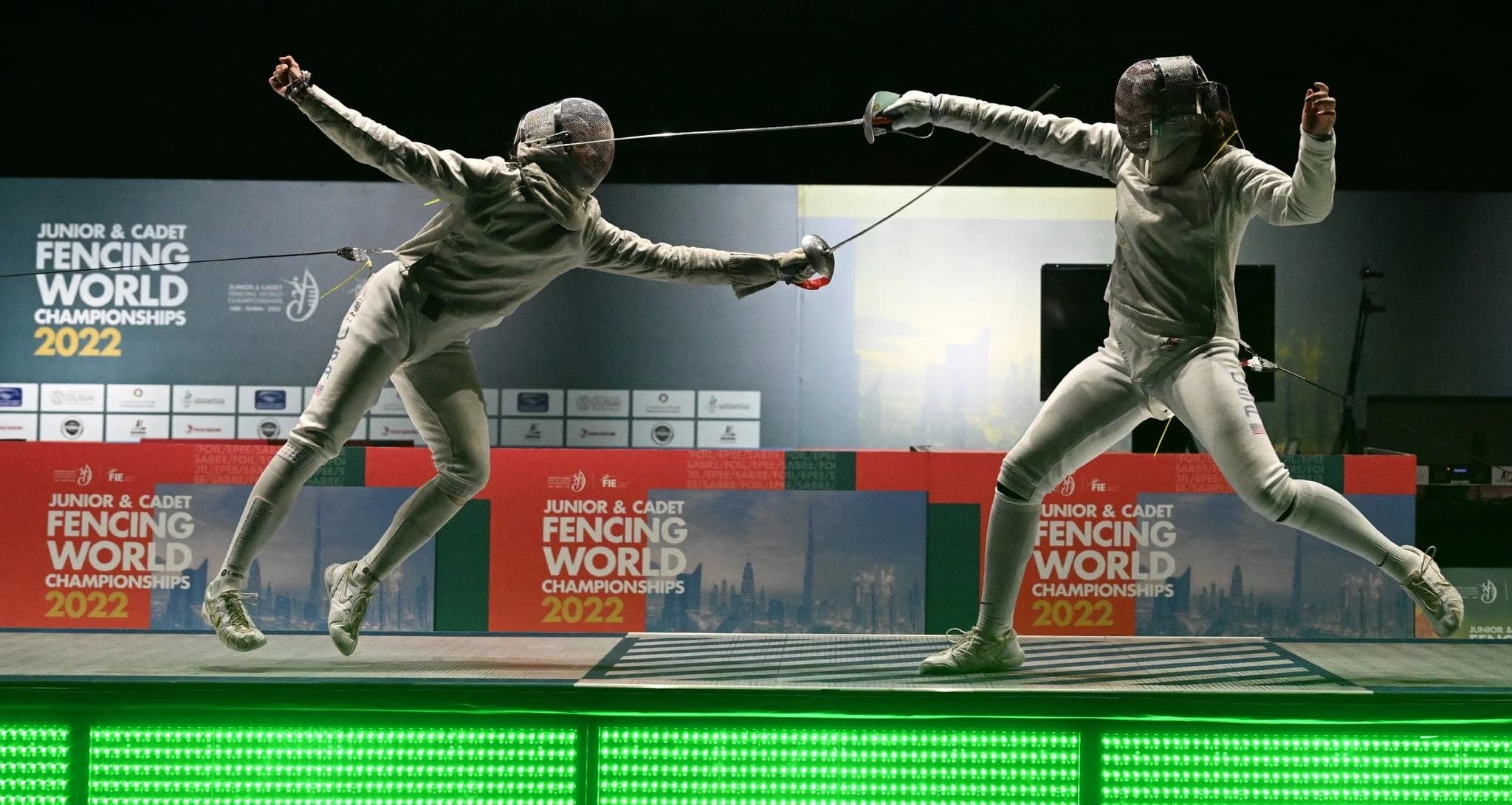 Two fencers in full protective gear lunge toward each other mid-bout during a match at the Junior & Cadet Fencing World Championships, captured in a dynamic moment of competition.
