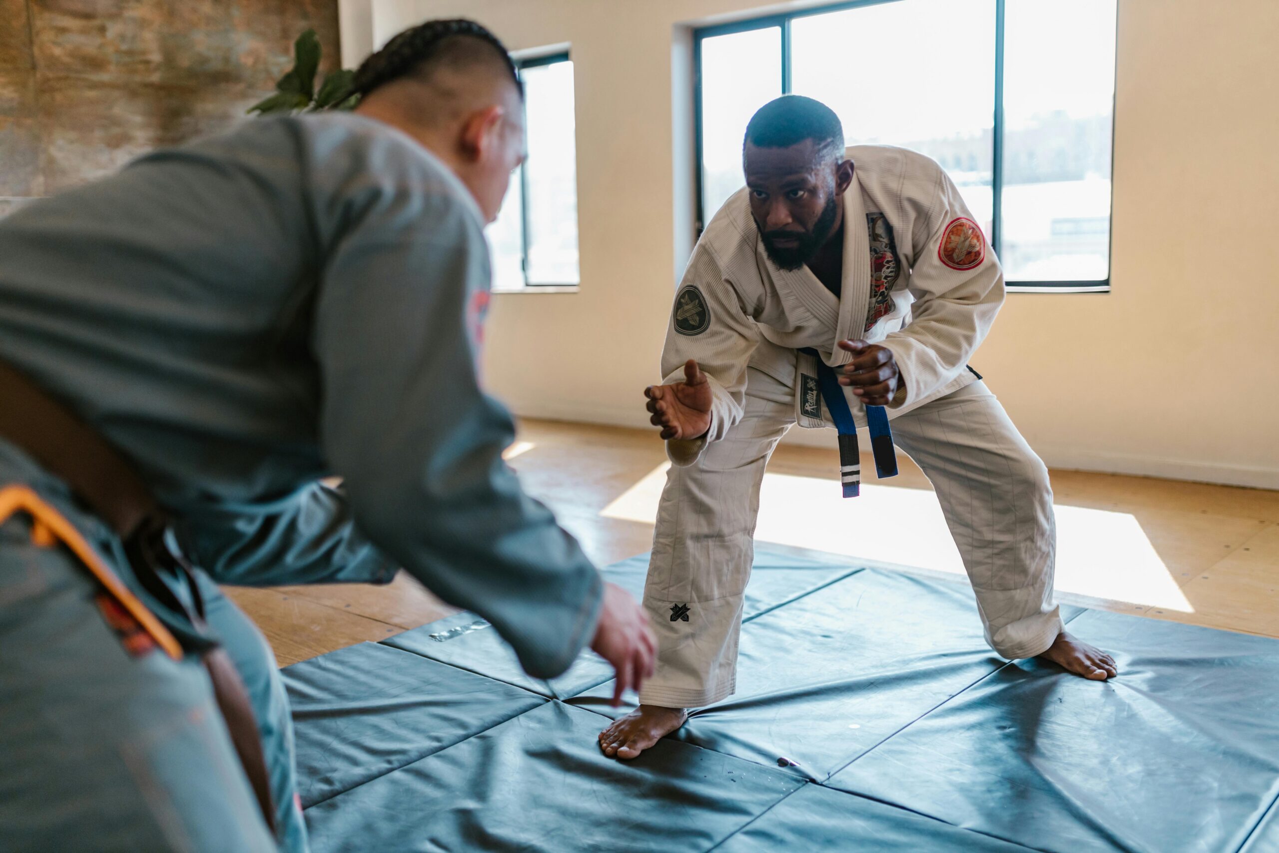 Two Brazilian jiu-jitsu practitioners face off on mats, crouched in a grappling stance during a training session in a bright studio.