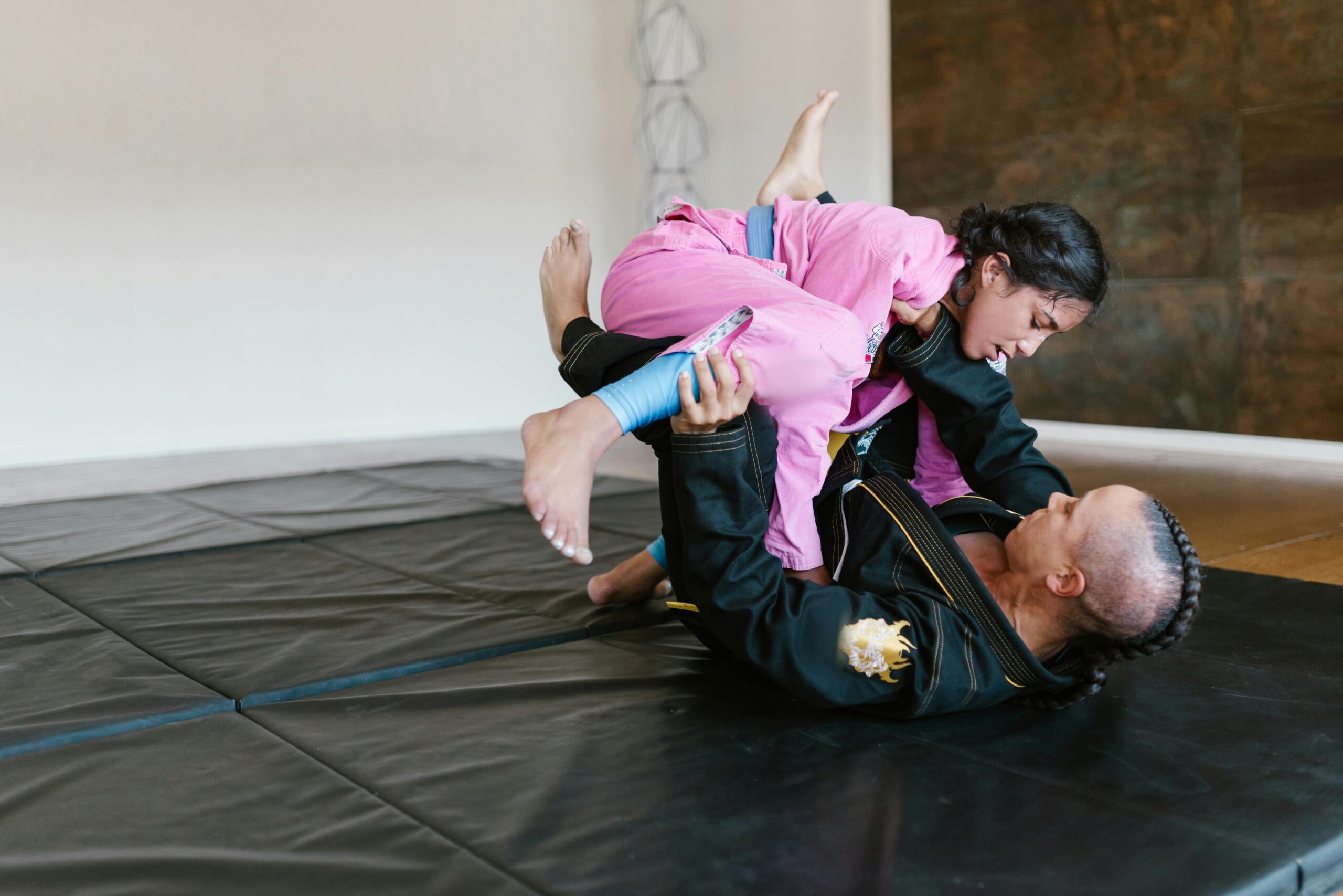 Two Brazilian jiu-jitsu practitioners spar on padded mats, with one athlete in a pink gi attempting to pass guard while the other in a black gi defends from the ground.