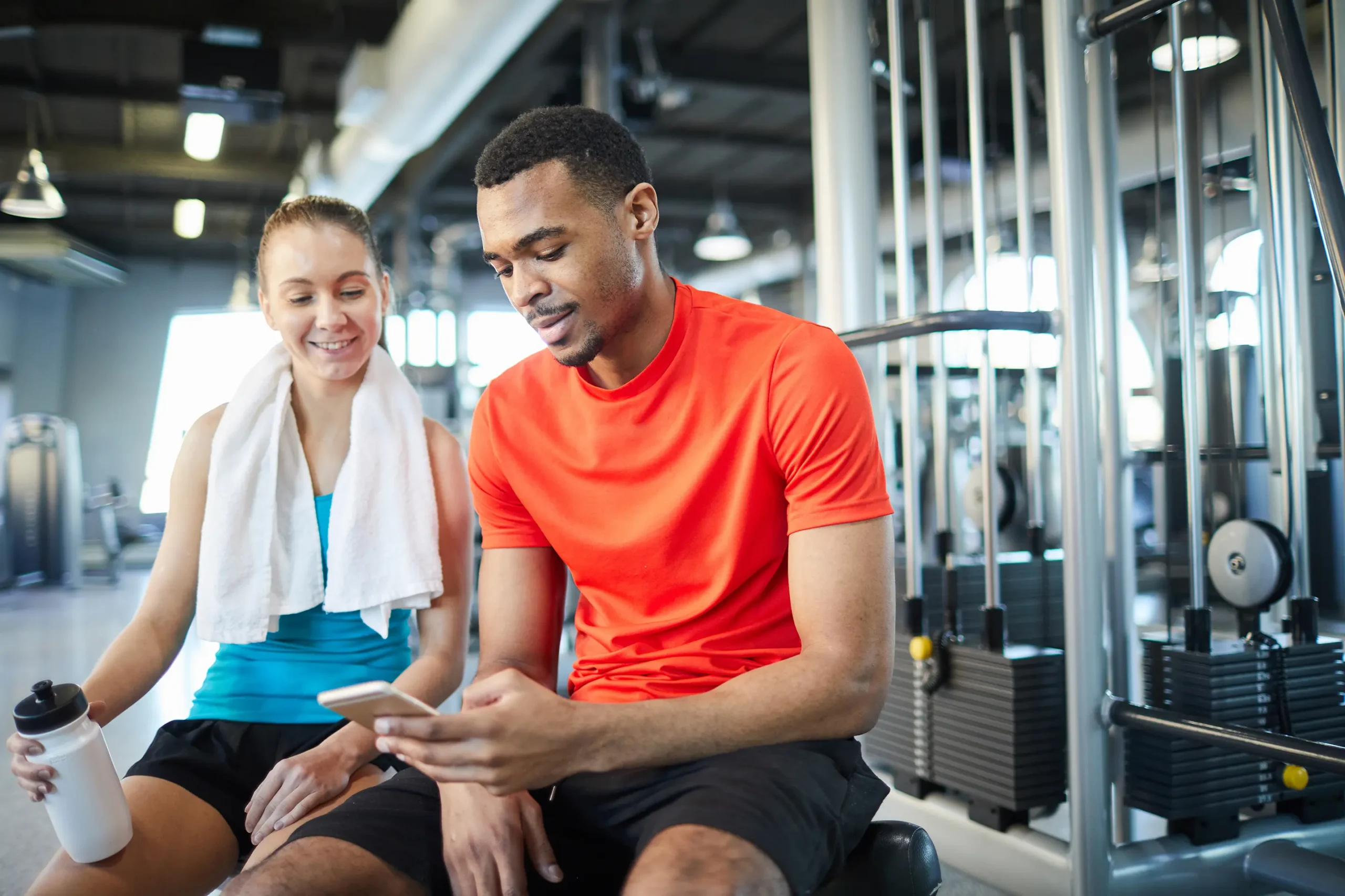 2a659dd4b0865606bdc07b7ce6a48151 Two people sit in a gym beside weight machines, smiling as they look at a smartphone after a workout.