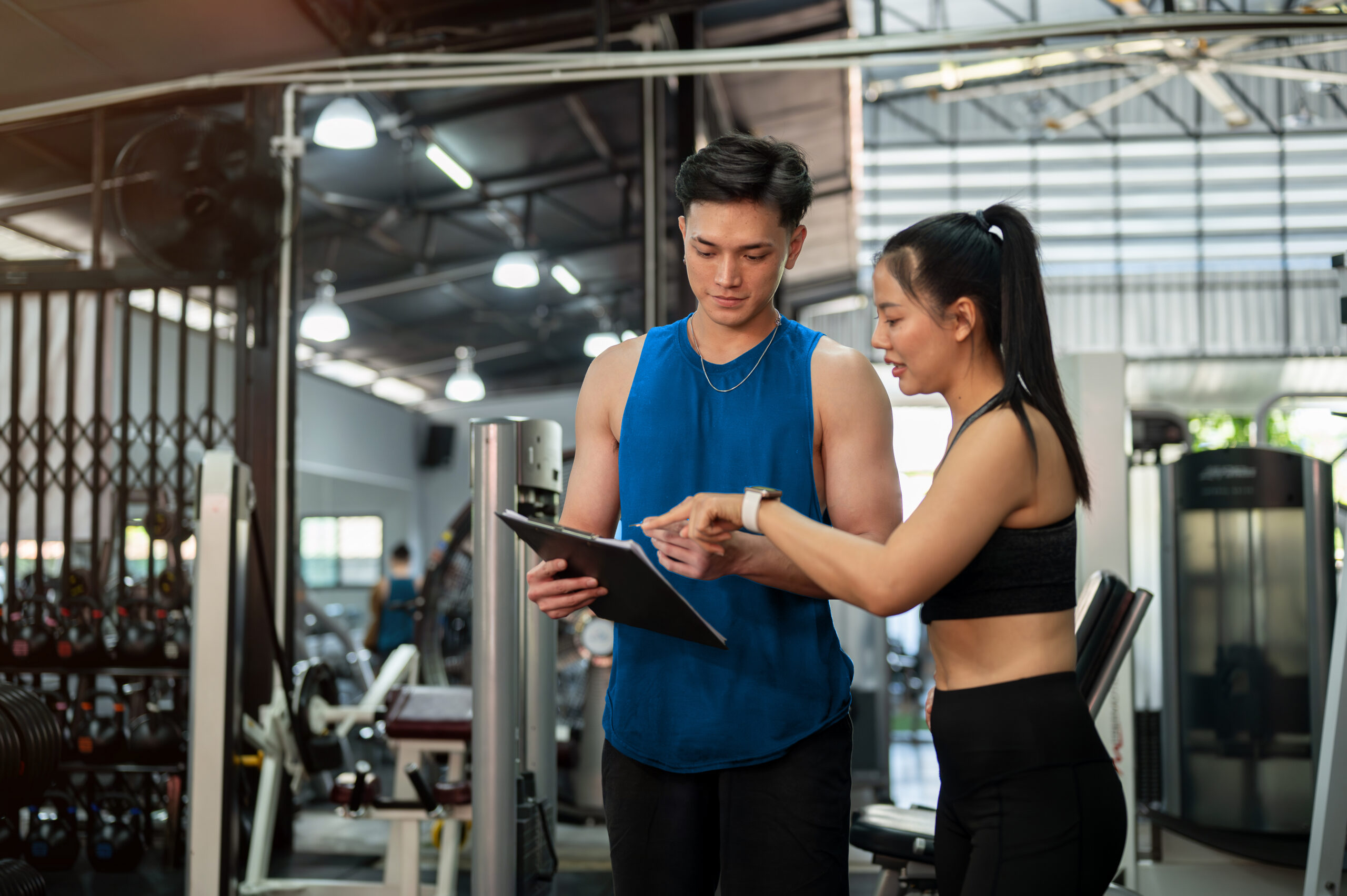 Personal trainer reviews a workout plan on a clipboard with a gym member while discussing fitness progress beside exercise equipment.