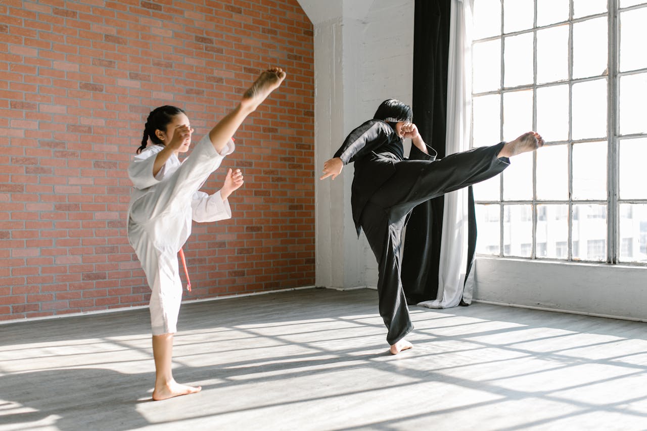 Child in a white martial arts uniform practices a high kick alongside an instructor dressed in black during a martial arts class in a bright training studio.