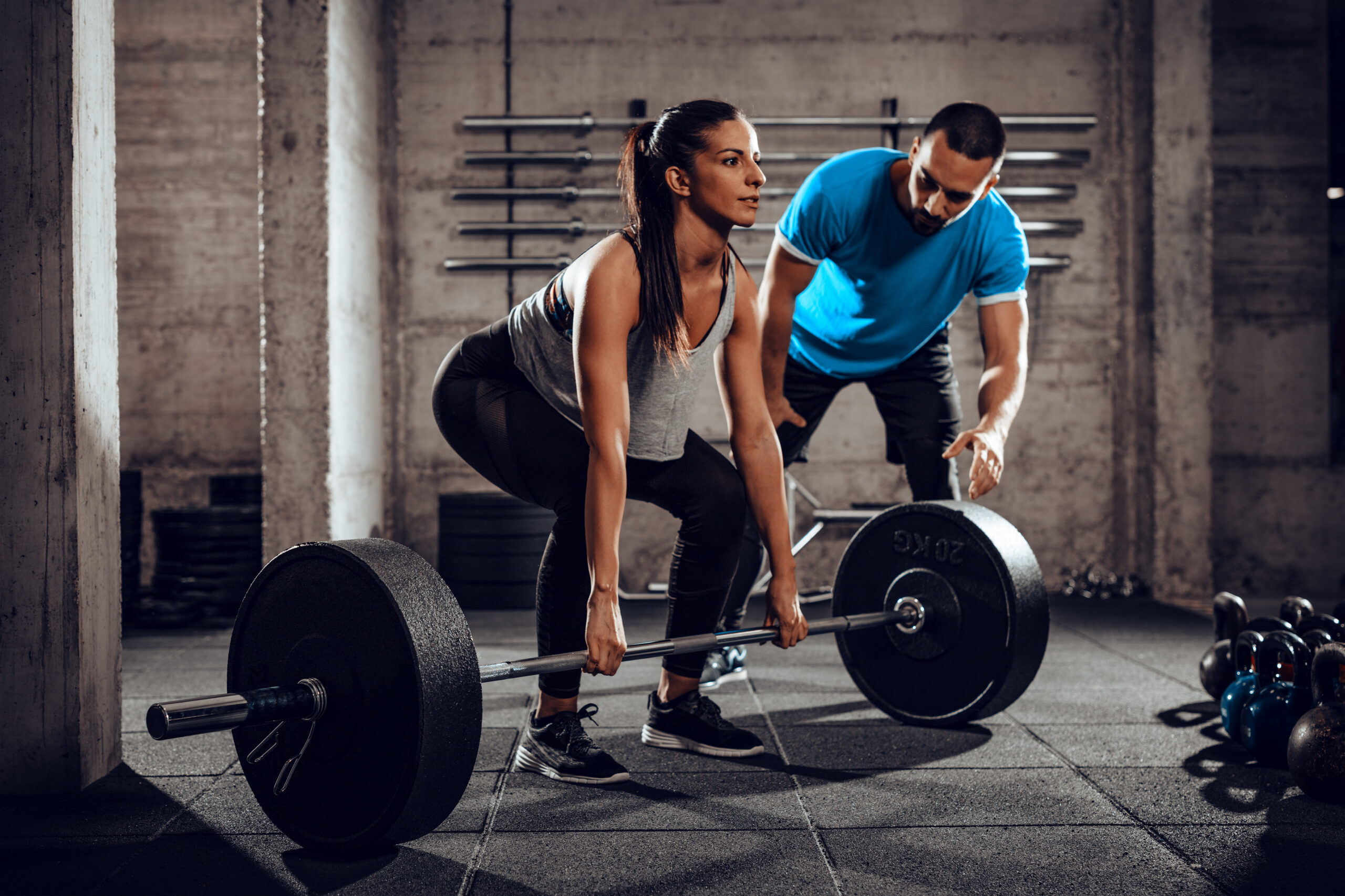 Personal trainer coaches a client through proper deadlift form during a barbell strength training session at a gym.