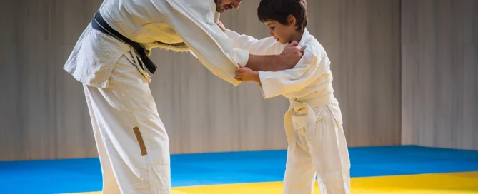 Martial arts instructor teaching a young student basic grappling techniques during a beginner class on training mats.