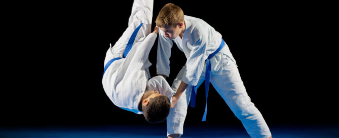 Boy performing a martial arts throw on another student during a judo training session, both wearing white uniforms with blue belts.