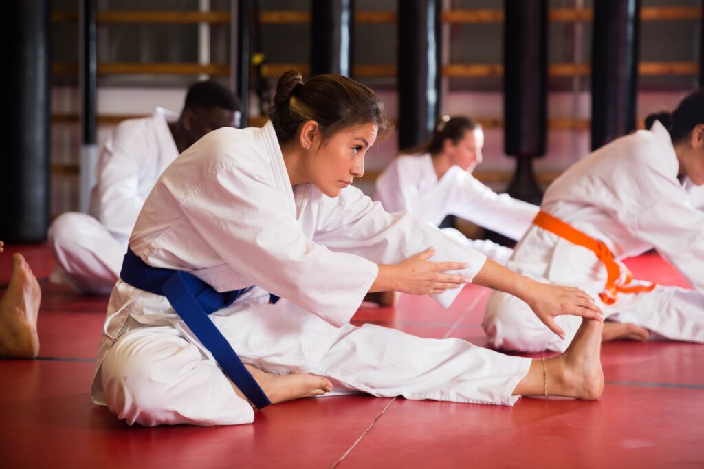 A group of karate students stretches on red training mats during class, with a student wearing a blue belt in the foreground reaching toward her foot while other students practice in the background.