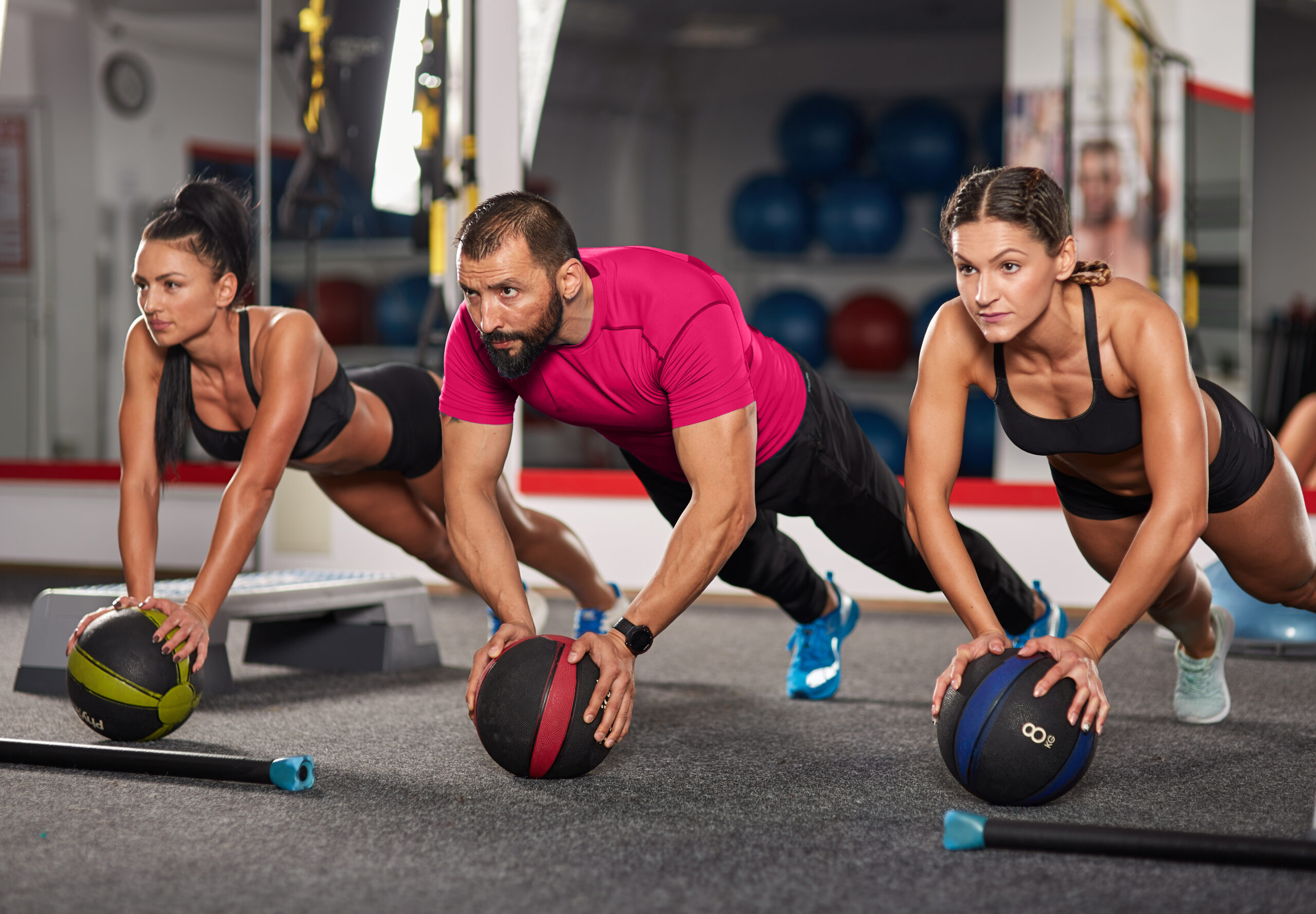 AdobeStock_181651811-vitality Three people perform plank exercises with medicine balls during a group functional fitness workout in a gym.