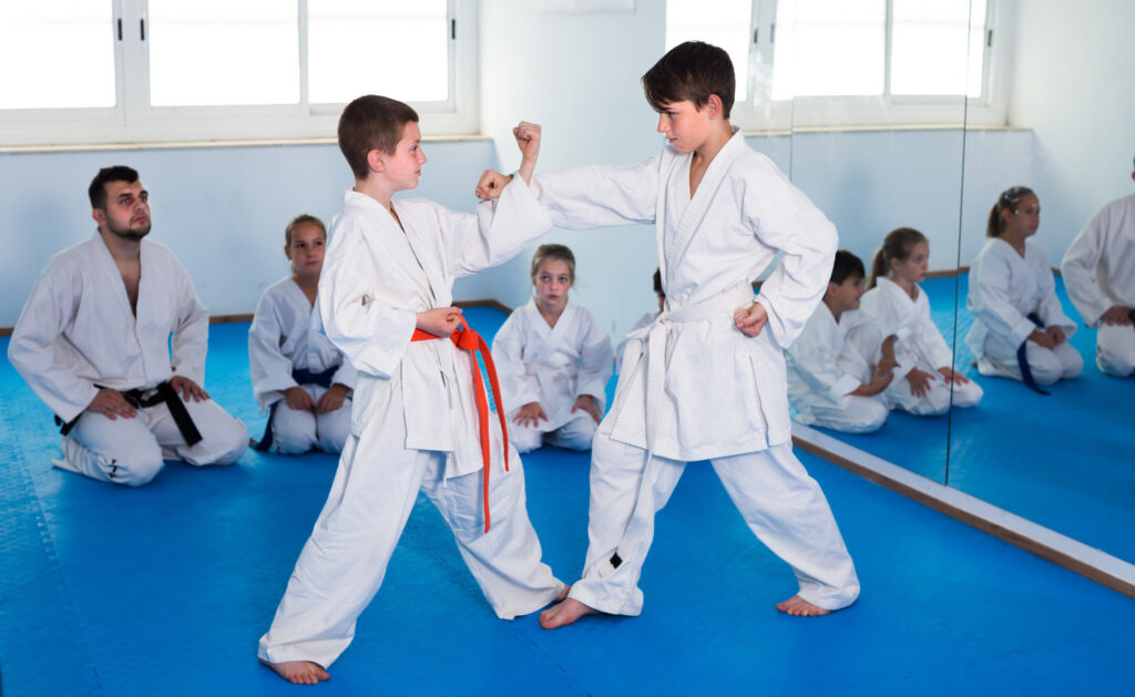 Friendly cheerful boys training in pair (1) Two young martial arts students practice blocking techniques during a supervised class on blue mats.