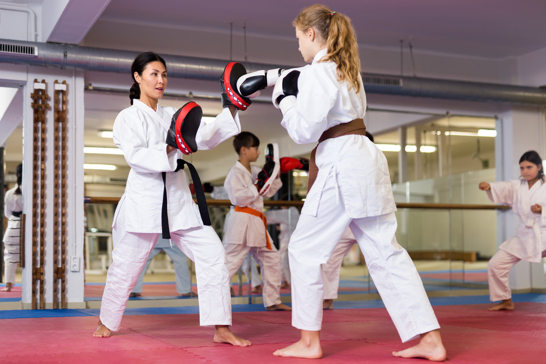 Young girls and boys in boxing gloves exercising jabs during group karate training. Woman trainer with focus mitts teaching girl in foreground (1) (1) Karate instructor holding focus mitts as a student practices punches during a martial arts class.