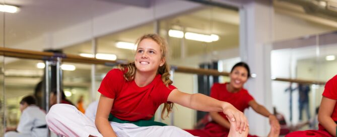 Young taekwondo students stretch on training mats during a warm-up session before class in a bright dojo.